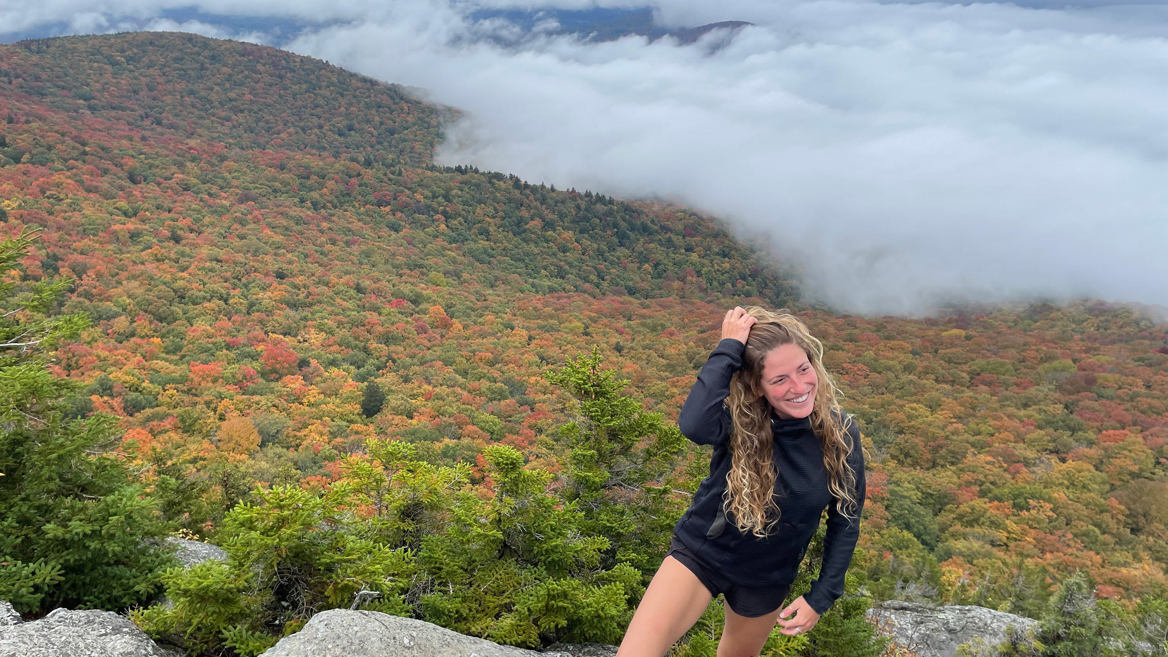 A woman poses on top of a mountain wearing a Black Thuja Burrow fleece hoodie. Autumn leaves and rolling clouds make the backdrop