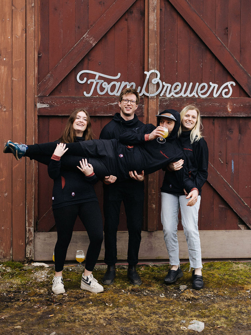 A group of Foam Brewers employees pose in front of a barn door sipping craft beer wearing Black Custom Thuja Burrow fleece hoodies with terracotta cuffs and chest embroidery.