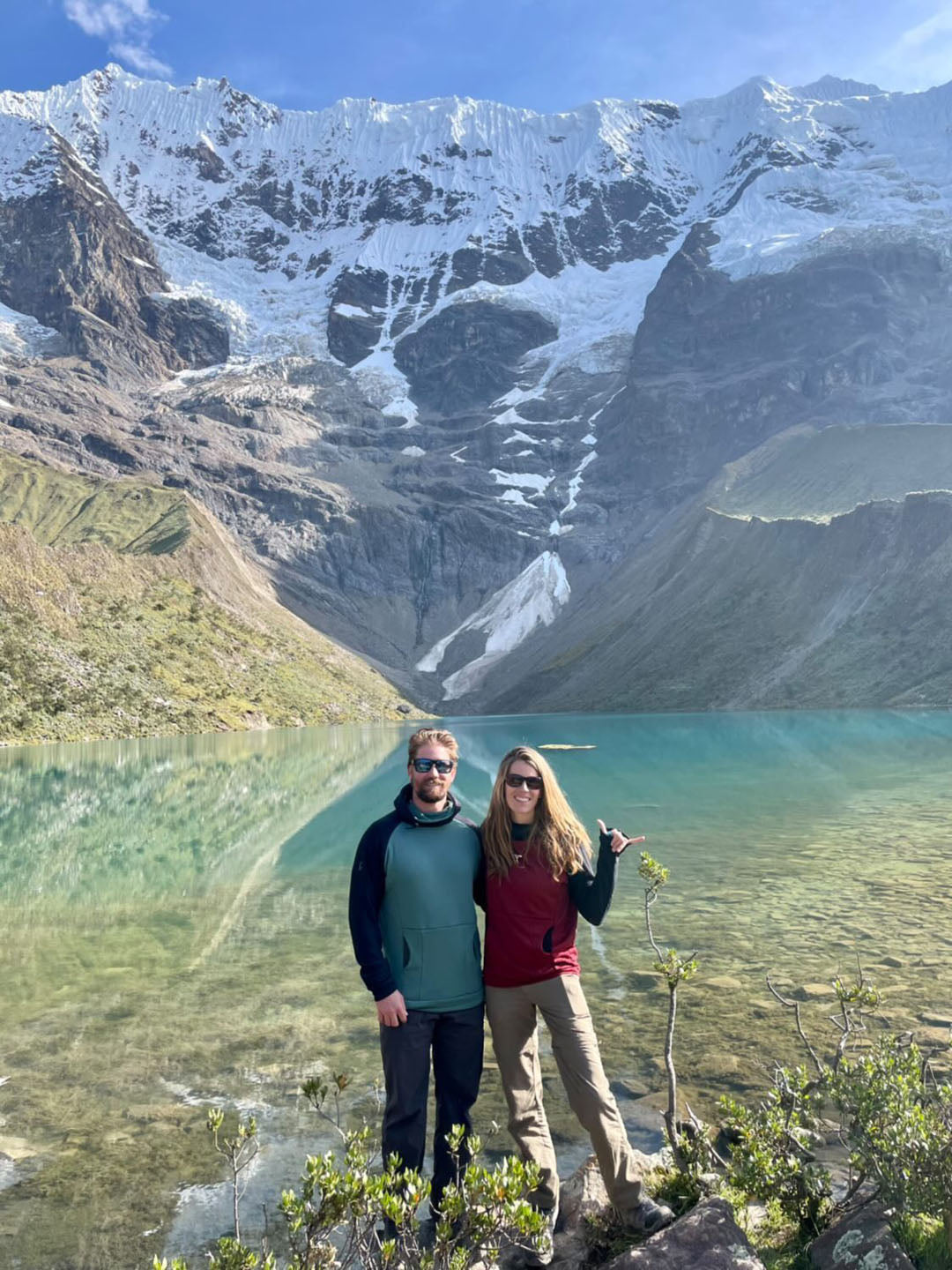 A woman wearing a Black and Classic Red custom Thuja Burrow fleece hoodie stands next to a man wearing a Black and Turquoise custom Thuja Burrow fleece hoodie in front of a crystal clear alpine lake with tall snow covered rocky mountains behind it.