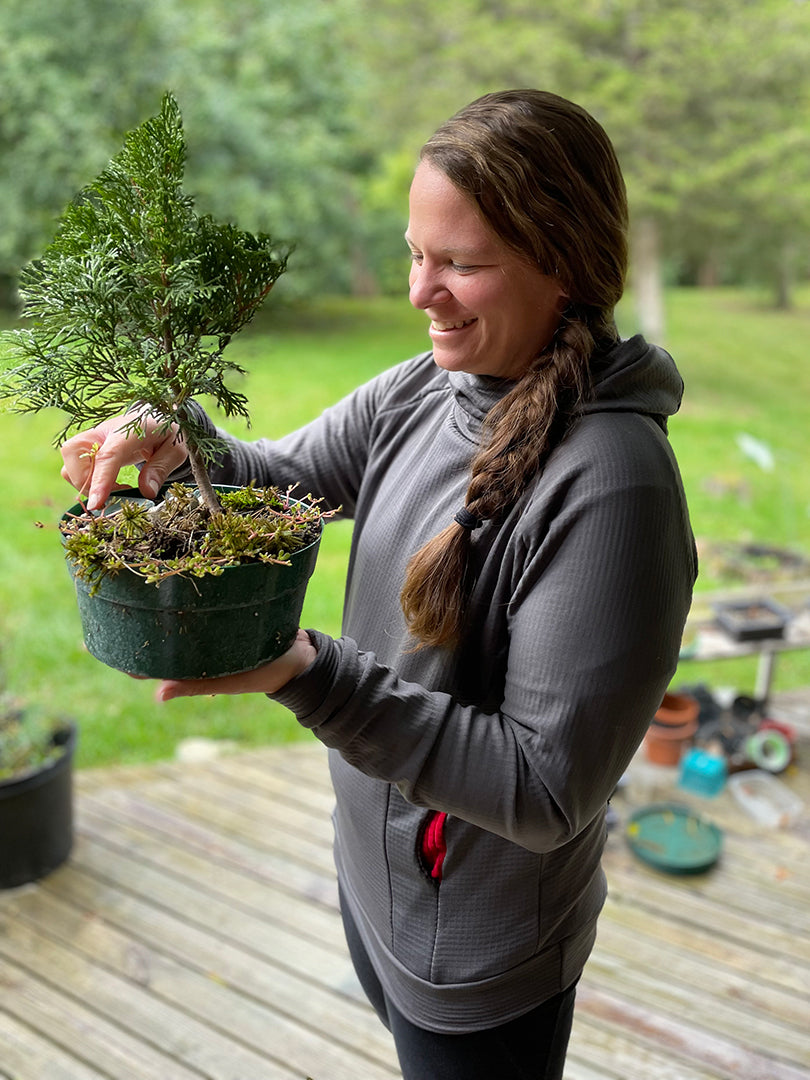 A woman wearing a Carbon custom Thuja Burrow fleece hoodie stands on rainy porch while inspecting a thuja bonsai she is holding.
