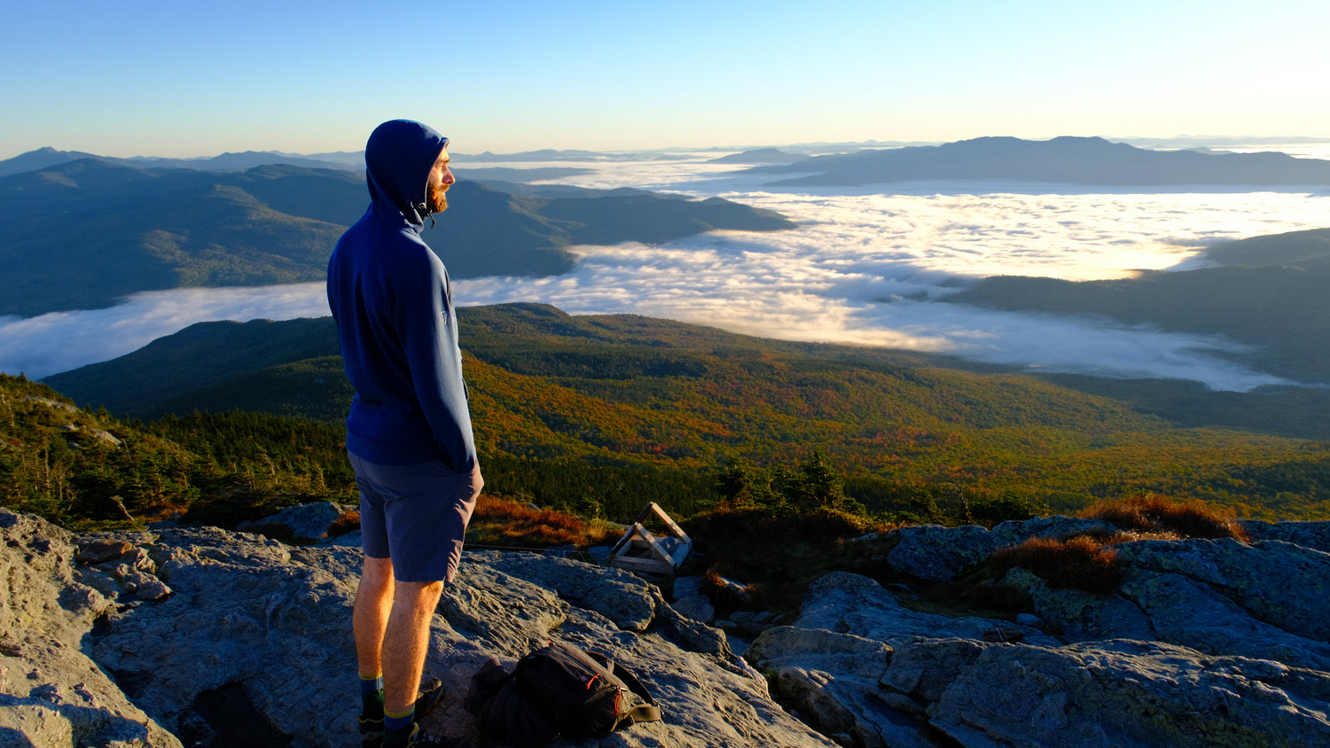A man stands on the top of a Vermont mountain wearing a Carbon Thuja Custom Burrow fleece hoodie looking a sea of inverted river valley fog and autumn foliage. 