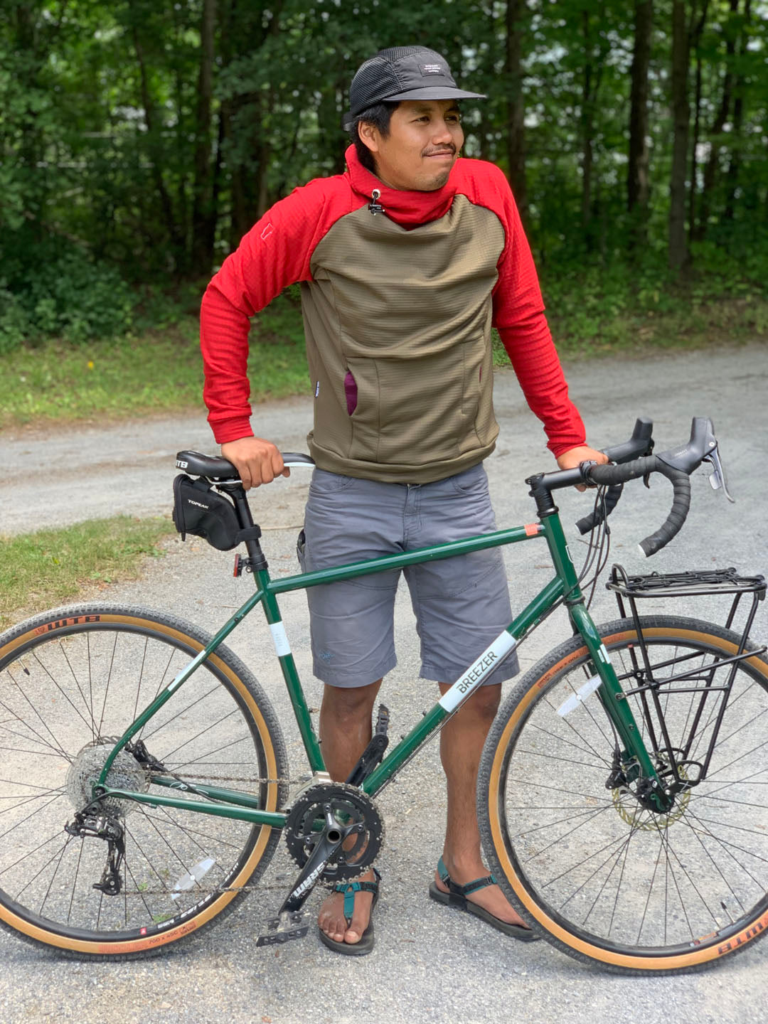 A man wearing a Classic Red and Olive custom Thuja Burrow fleece hoodie stands on a gravel driveway holding his Breezer bicycle in front of him.