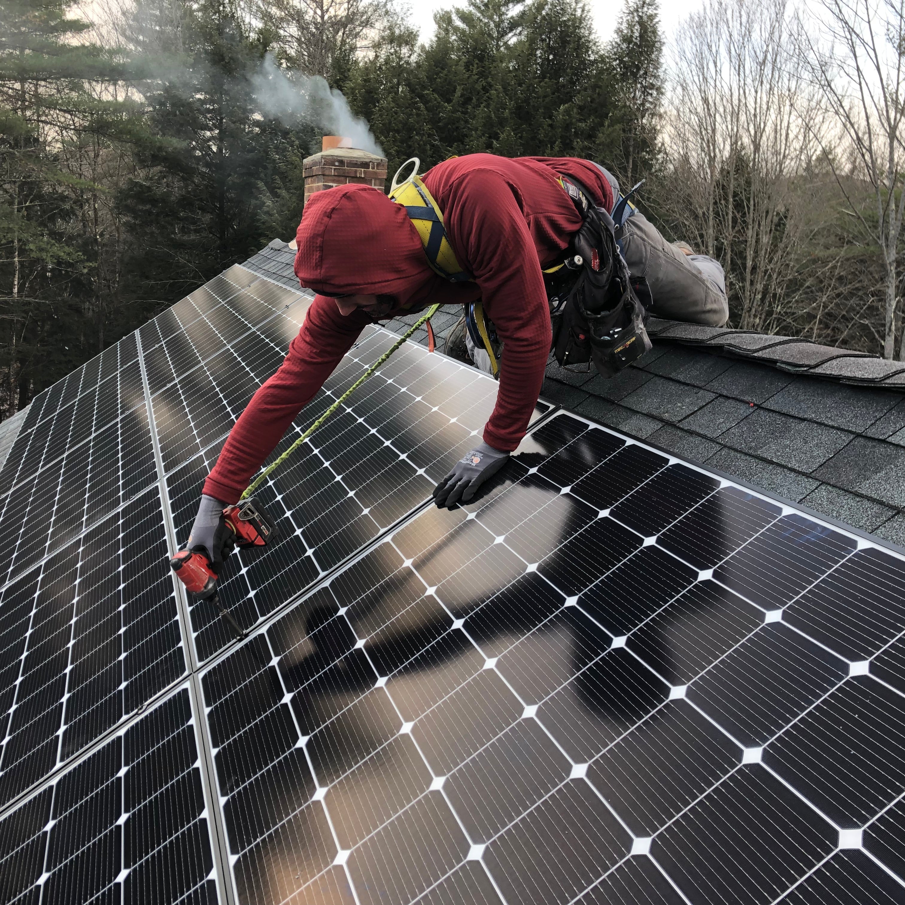 A man wearing a Maroon custom Thuja Burrow fleece hoodie with the hood on leans over the peak of a roof while installing solar panels.