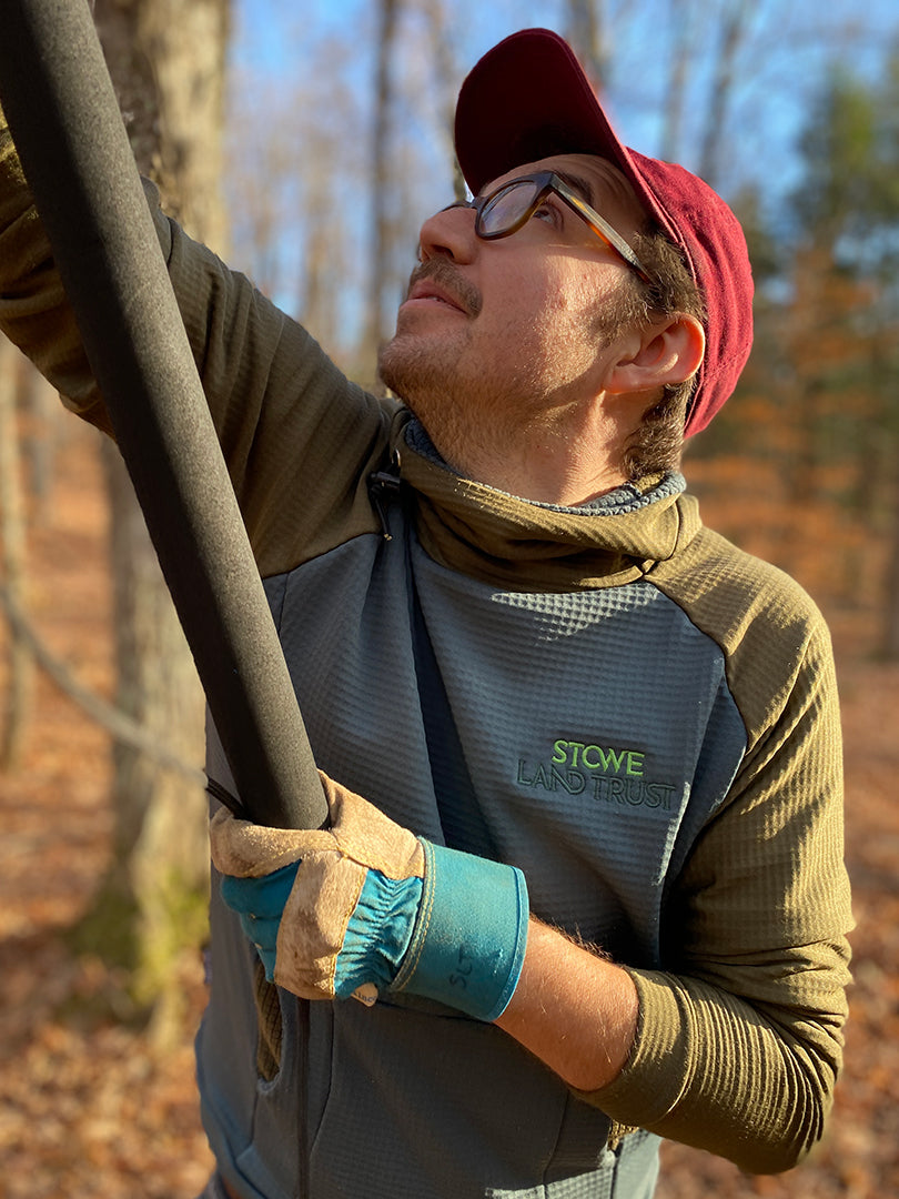 A member of Vermont's Stowe Land Trust pruning a tree in autumn wearing a Custom Thuja Burrow fleece Hoodie in Olive and Slate with "Stowe Land Trust" embroidered on the chest.