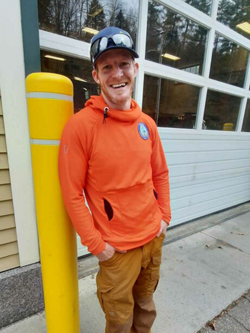 A member of Stowe Mountain Rescue standing in front of the Stowe Vermont Fire Department garage door wearing an orange Custom Thuja Burrow fleece Hoodie with an attached Stowe Mountain Rescue Patch on the chest.  