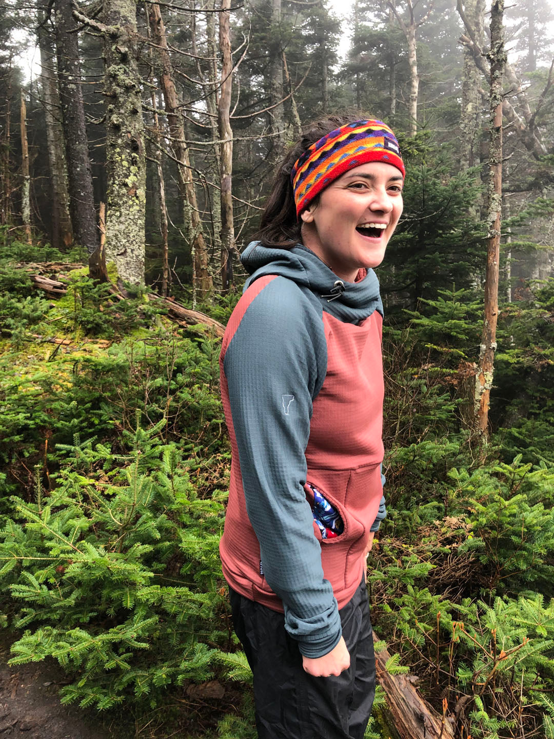 A woman wearing a Slate and Terracotta custom Thuja Burrow fleece hoodie laughs while hiking through a rainy and foggy conifer laden trail.