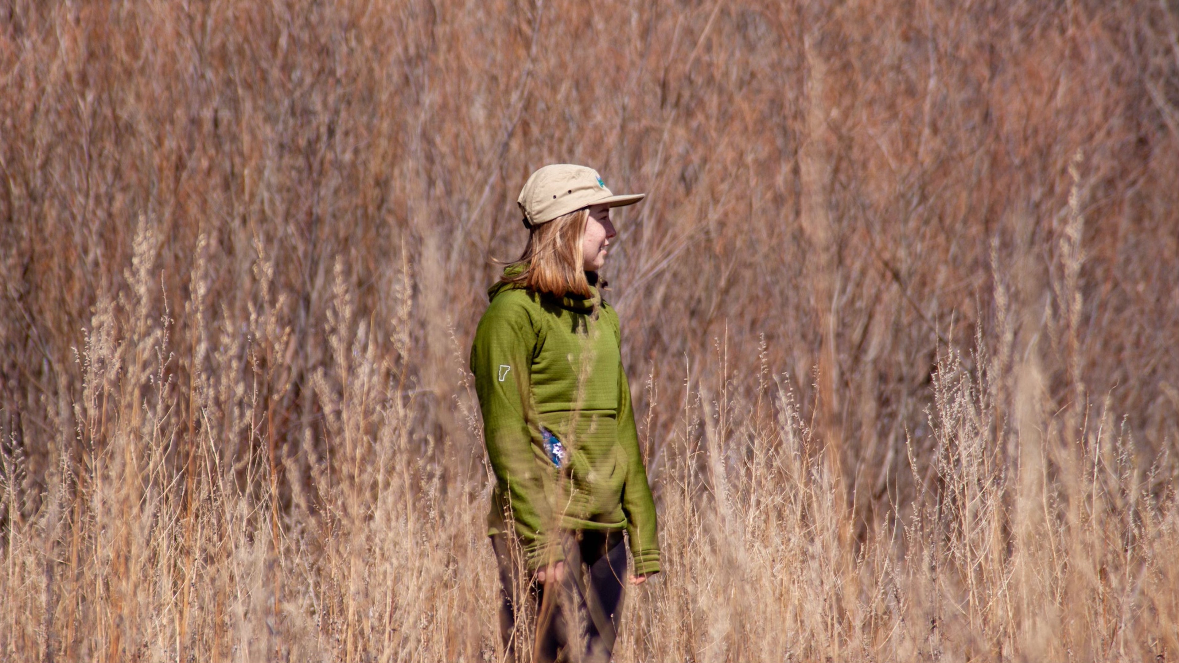A woman stands on a field edge in dried autumn grass wearing a custom Sprout Green Thuja Burrow fleece hoodie.