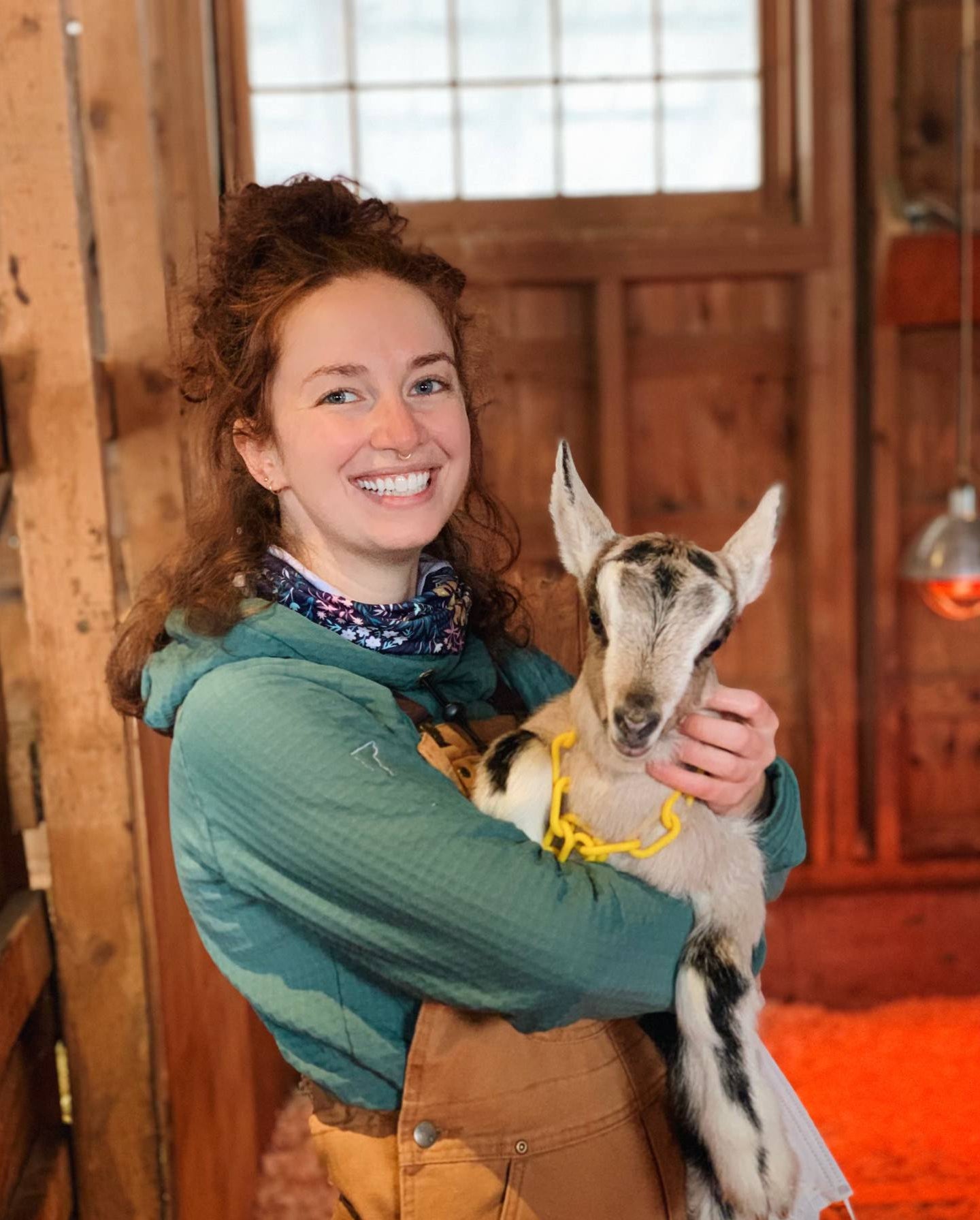 A woman wearing a Teal custom Thuja Burrow fleece hoodie holds a baby goat kid inside of wooden barn.