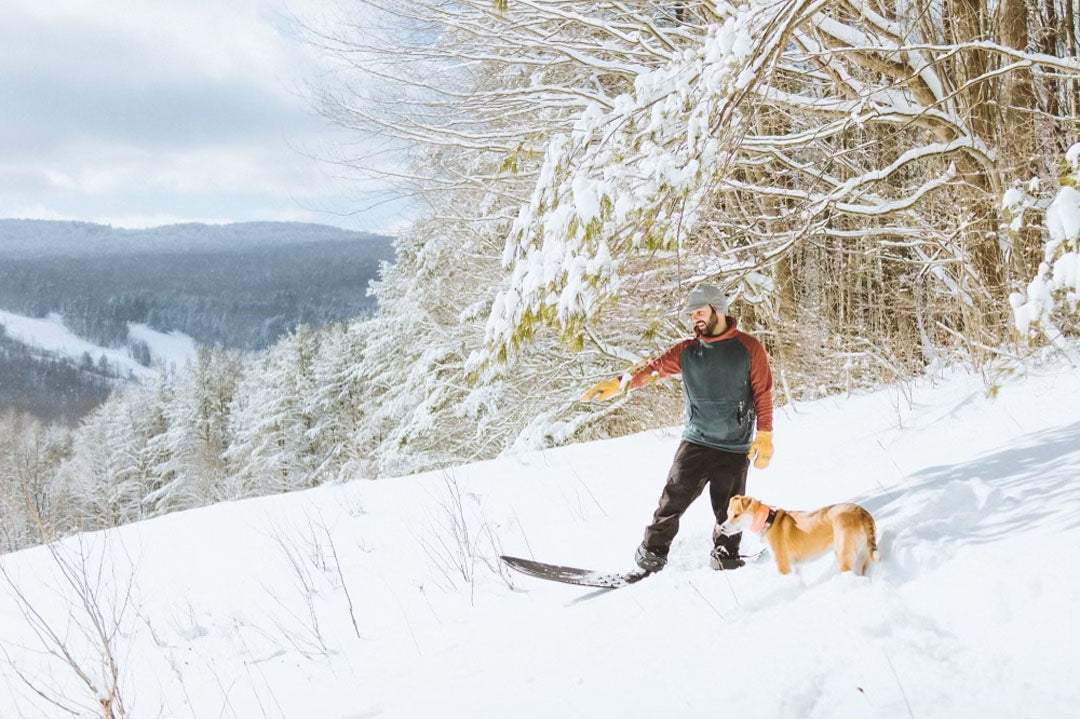 A man on a snow snurfer wearing a Terracotta and Slate Thuja custom Burrow fleece hoodie points down a powdery snowy hill with his dog. 