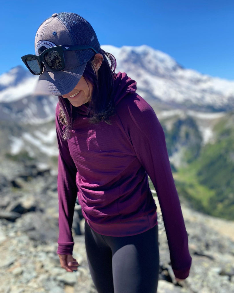 A woman in a Plum Thuja Ultralight Hell Brook Hoodie steps forward on a high altitude mountain with melting snow and conifers in the background.
