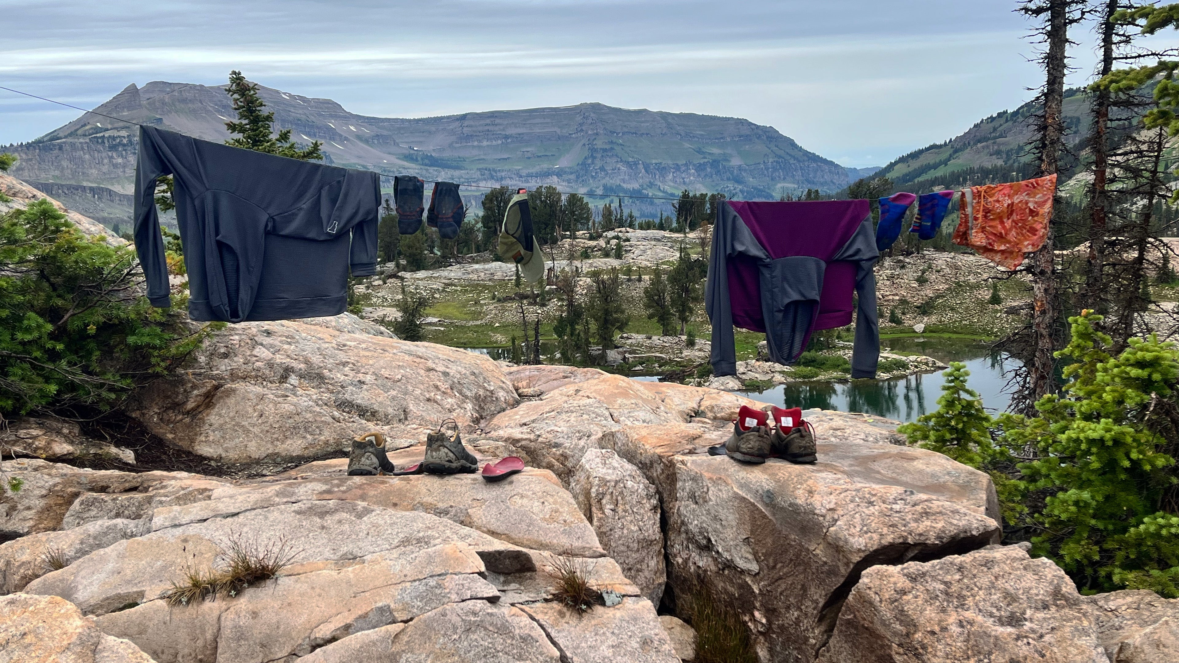 Two Thuja Ultralight Hell Brook Hoodies, one Forge Grey and one Forge Grey Plum combination, hanging on a clothes line in the backcountry mountains of Grand Teton National Park. 