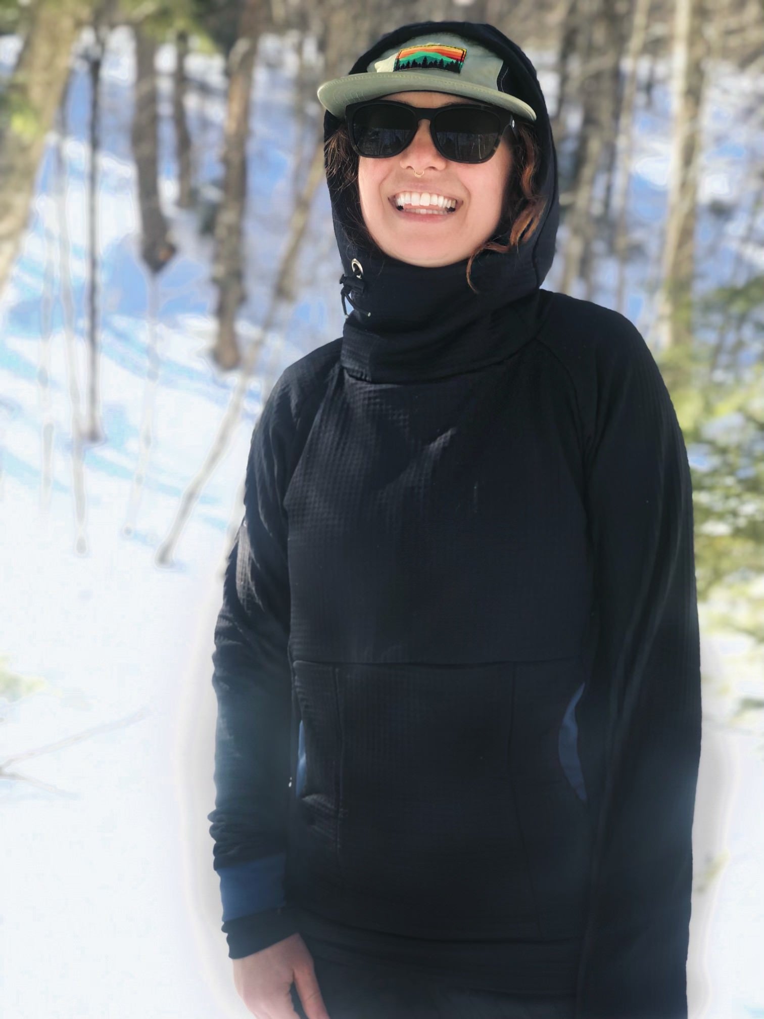A woman wearing a Black Thuja Burrow fleece hoodie with the hood up smiles in a sunny, snowy, forest.