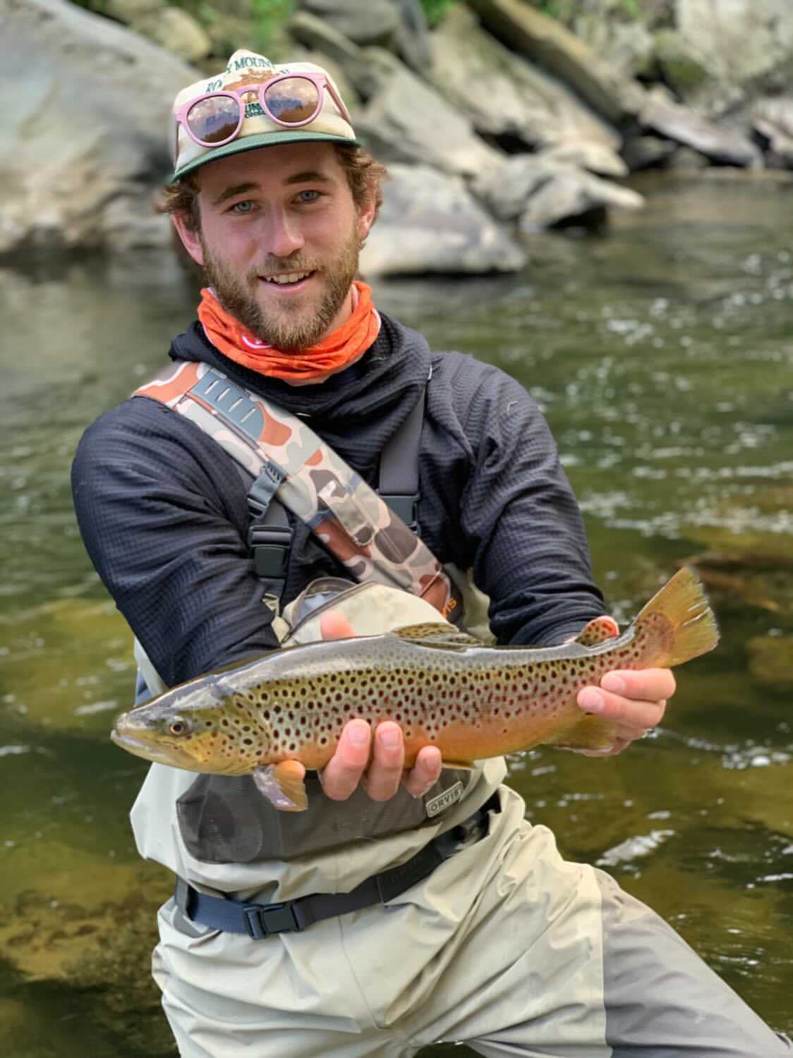 A man wearing a Black Thuja Burrow fleece hoodie, waders and other fishing gear extends a freshly caught brown trout while kneeling in a clear, rocky river.