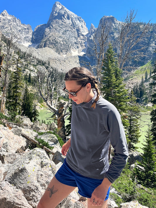 A woman wearing a Forge Grey Thuja Ultralight Hell Brook hoodie hikes up a boulder field in the Grand Tetons.